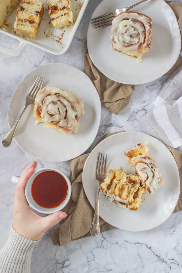 Flatlay showing chelsea buns on 3 plates with a hand holding a cup of rooibos tea