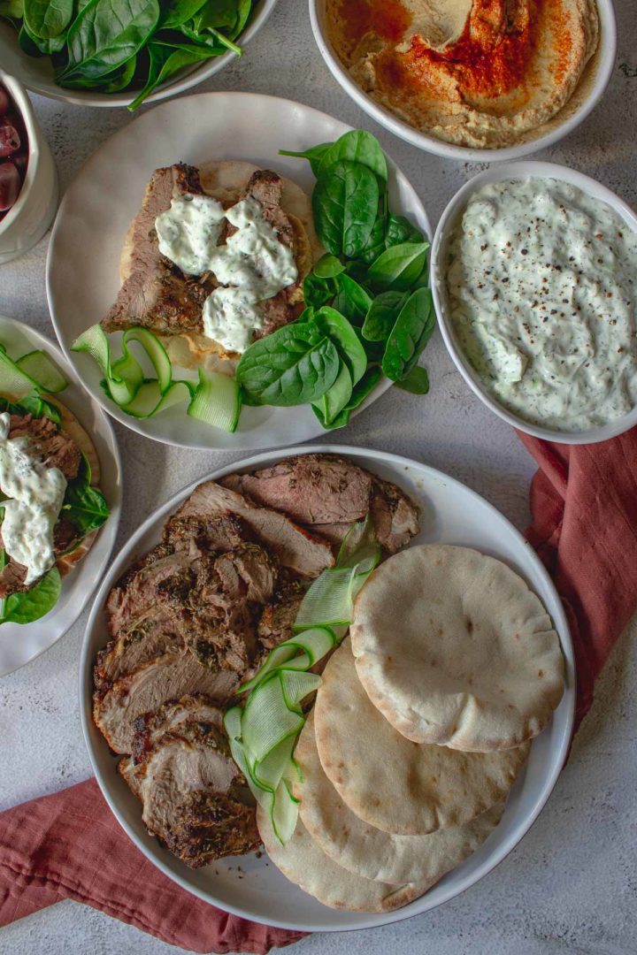 roast leg of lamb carved and plated next to pitas with tzatziki, hummus and spinach