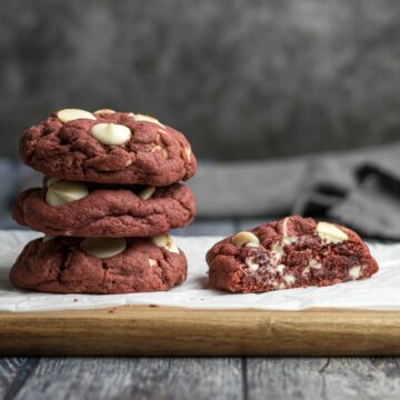 Three red velvet cookies stacked on each other next to a red velvet cookie sliced in half to show the cross section and white chocolate chips.