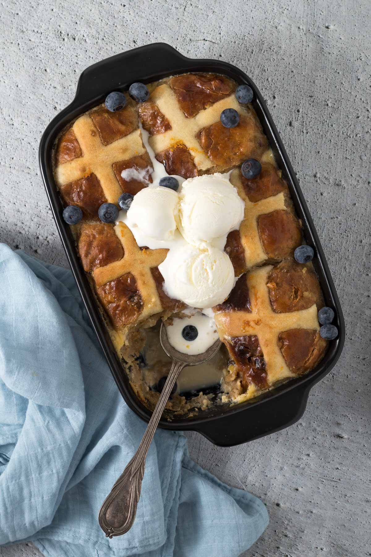 Hot cross bun pudding in serving dish with vanilla ice cream and fresh blueberries.
