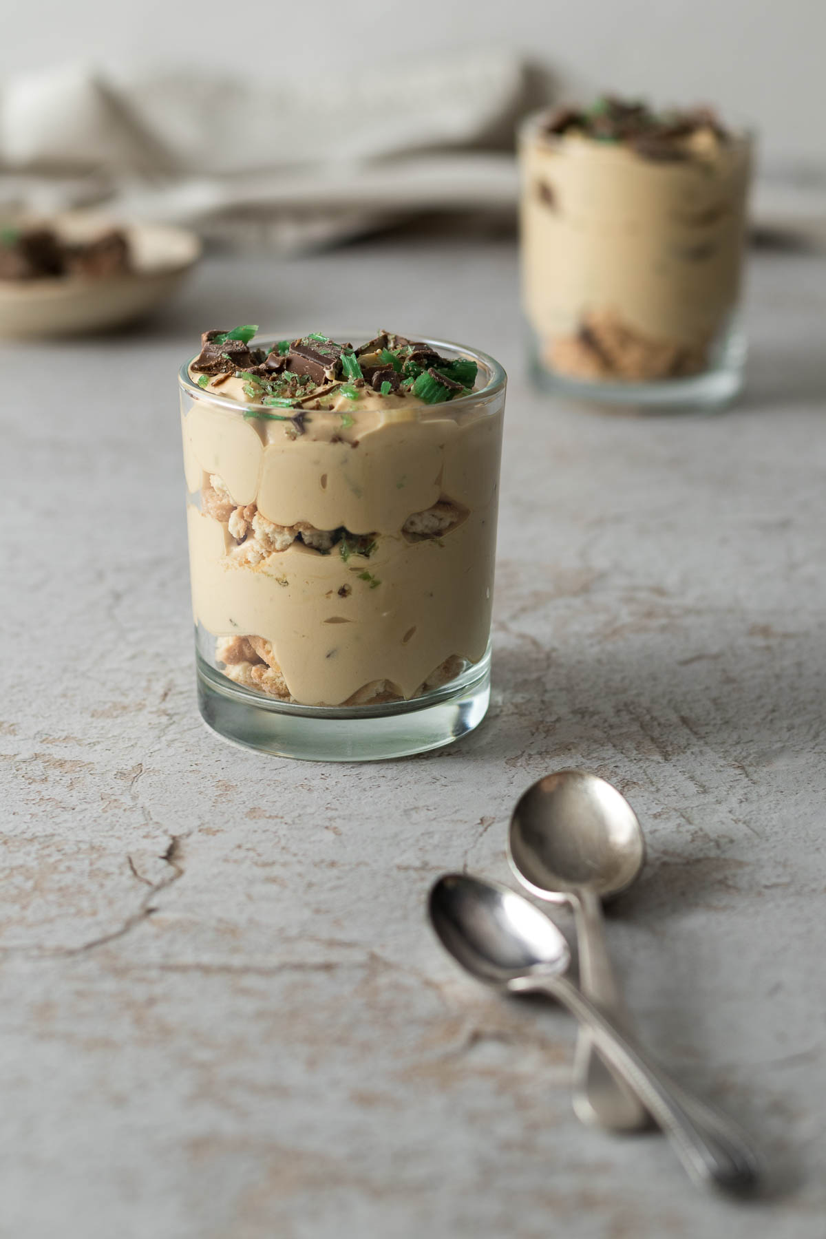 An individual portion of the peppermint crisp tart in a glass showing the layers of biscuit, caramel-cream and mint chocolate pieces.